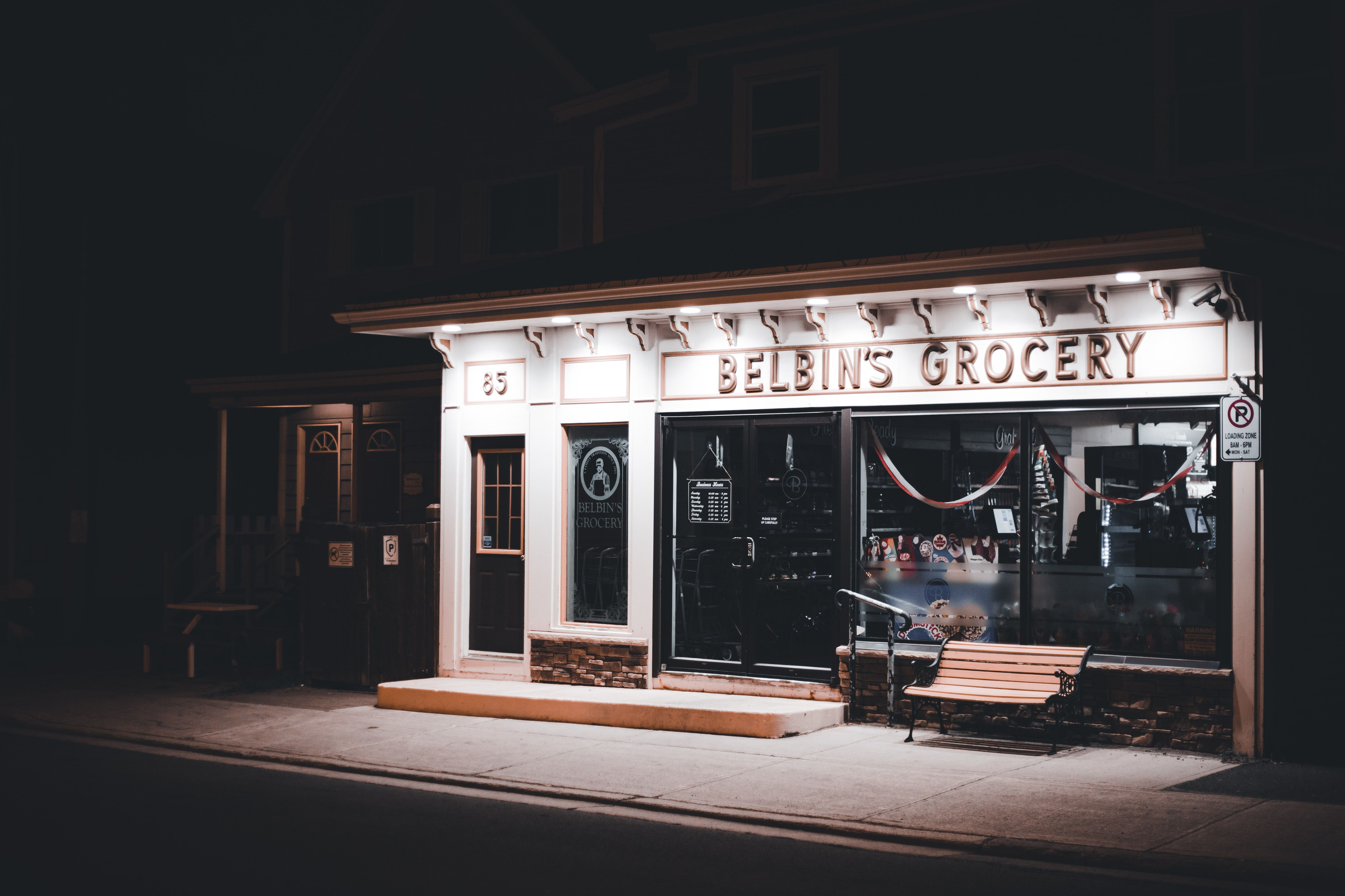 Grocery storefront glowing with warm exterior lighting at night, with a bench out front